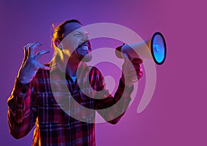 Portrait of angry emotional man shouting in megaphone with annoyance against purple studio background in neon light