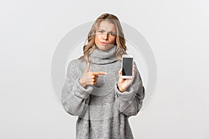 Portrait of angry girl pointing finger at smartphone, showing something disappointing and waiting for explanation