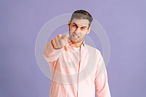 Portrait of angry handsome young man pointing index finger at you on purple  background, looking at camera.