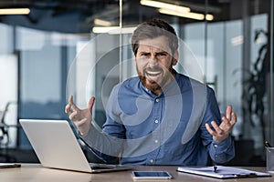 Portrait of an angry and irritated young businessman sitting at a desk in the office and shouting at the camera