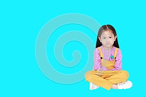 Portrait of angry little Asian child girl in pink-yellow pink-yellow dungarees sitting isolated on cyan background with copy space