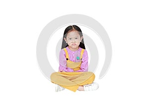 Portrait of angry little Asian child girl in pink-yellow pink-yellow dungarees sitting isolated on white background with copy