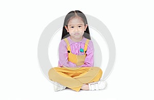 Portrait of angry little Asian child girl in pink-yellow pink-yellow dungarees sitting on white background