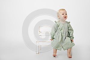 Portrait of angry little girl with blue eyes wearing green cotton dress, sit on white wooden little bench, grimacing.