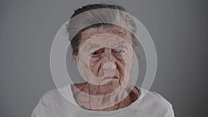Portrait of an angry senior Caucasian woman with gray hair and deep wrinkles looking at camera on gray background. Very