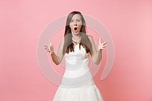 Portrait of angry shocked bride woman with opened mouth in white wedding dress standing spreading hands isolated on pink