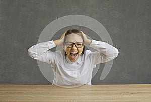 Portrait of angry stressed young woman sitting at desk, holding her head and screaming