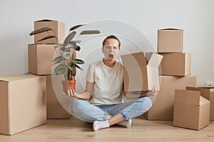Portrait of angry woman wearing white T-shirt sitting on the floor near cardboard boxes with pile, holding flower pot with plant