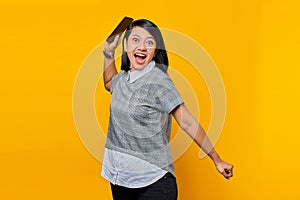 Portrait of angry young Asian woman with displeased expression and holding mobile phone over yellow background