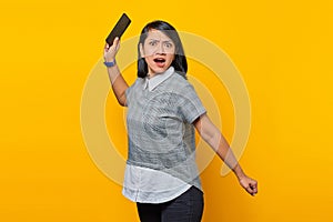 Portrait of angry young Asian woman with displeased expression and holding mobile phone over yellow background