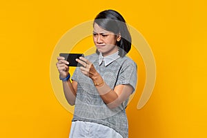 Portrait of angry young Asian woman playing video game on mobile phone over yellow background