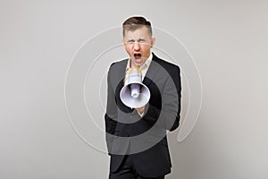 Portrait of angry young business man in classic black suit and shirt screaming on megaphone isolated on grey wall