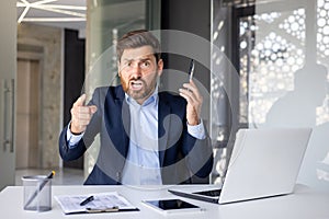 Portrait of an angry young man businessman sitting in the office at the table, talking on the phone, shouting and