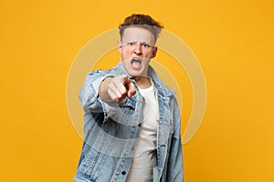 Portrait of angry young man in denim casual clothes pointing index finger on camera, swearing isolated on yellow orange