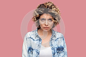 Portrait of angry young woman with curly hairstyle in casual blue shirt standing and looking at camera with agressive temper face