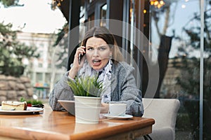 Portrait of angry young woman solving business problems shouting during phone conversation sitting in cafe terrace