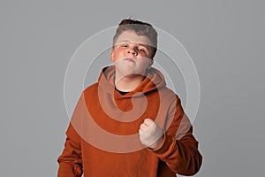 Portrait of annoyed angry plus size teenager boy shaking fist, standing over gray isolated background, looking at the camera