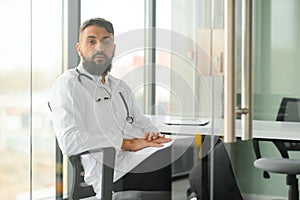 Portrait of arabic doctor handsome young man in workwear posing at modern clinic, sitting at workdesk