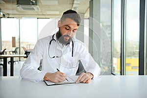 Portrait of arabic doctor handsome young man in workwear posing at modern clinic, sitting at workdesk