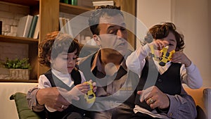 Portrait of arabic father sitting on sofa with his twin sons playing with toy-robots in living room.