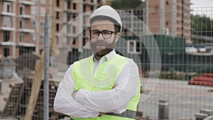 Portrait of architect man standing on the construction site with crossed hands looking at the camera. Outside, slow