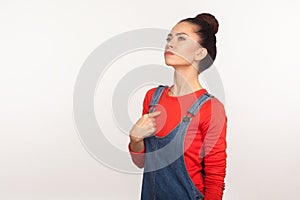 Portrait of arrogant egoistic girl with hair bun in denim overalls pointing herself and looking up with proud self-confident
