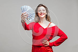 Portrait of arrogant wealthy beautiful woman in red dress holding money, isolated on gray background