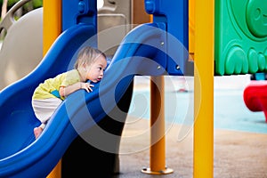 Portrait of an Asian baby boy seriously trying to climb blue slide, Toddler playing with toy amidst an outdoor playground.