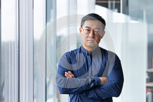 Portrait of asian businessman inside modern office, boss in glasses serious looking at camera, man with crossed arms