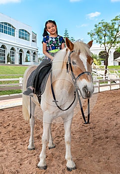 Portrait of Asian child girl riding on a white horse, having face mask hangs on her neck, full-body of child riding horse, travel