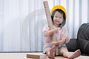 Portrait of Asian child girl wearing yellow helmet safety hat. Kid holding paper cylinder for work. Pretend to be an engineer.