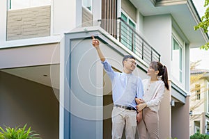 Portrait of Asian couple walking hugging and pointing together looking happy in front of their new house to start new life. Family