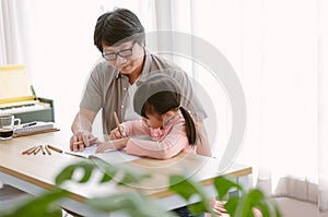 Portrait of Asian father and cute little daughter sitting at table drawing a picture or writing book with colour pencils at home.