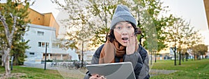 Portrait of asian girl with shocked, surprised face, holds tablet, read or watched big news, looks amazed, sits in park