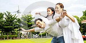 Portrait of asian happy family of three having fun together in park, little daughter sitting on father back and mother smile