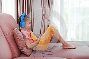 Portrait Asian little girl child wearing headphone and using smartphone on sofa in living room at home