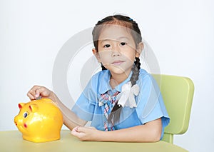 Portrait of Asian little girl in school uniform sitting on table and putting coin into piggy bank isolated on white background.