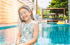 Portrait Asian little girl at swimming pool, casual cute dress, smiling face, blurred background of swimming pool at a resort