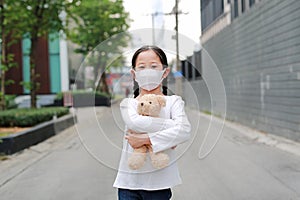 Portrait asian little girl wearing mask to protect corona virus and air pollution pm2.5 dust with hugging teddy bear doll standing