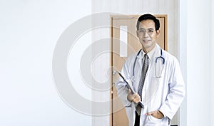 Portrait of asian male doctor at the hospital ward in front of patient room holding clipboard with medical record for better