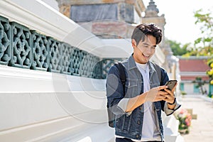 Portrait of Asian male traveler using a mobile phone on sidewalk of buddhist temple on street in Bangkok, Thailand