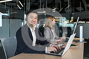 portrait of Asian man in business attire, call center employee looks at camera and smiles, businessman uses computer for