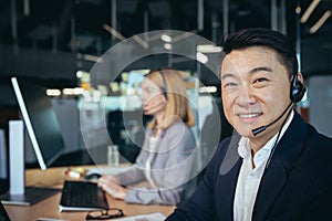 Portrait of Asian man in business attire, call center employee looks at camera and smiles, businessman uses computer for