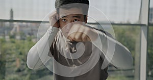 Portrait of Asian man fighter punching with fists and looking at camera training in glass wall gym