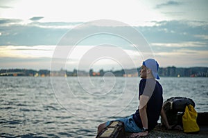 Portrait Asian man sitting on the port and looking out to the sea with the twilight sky of sunset and bokeh lights of the city in