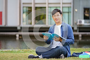 Portrait of Asian man university student aitting on grass in campus looking at camera and smile and reading a book in park