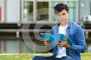 Portrait of Asian man university student aitting on grass in campus looking happy and reading a book in park