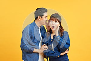 Portrait of asian man whispering secret or interesting gossip to excited woman in her ear isolated over yellow background