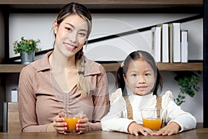 Portrait of Asian mother and daughter girl child drinking fresh orange juice during sitting in living room at home, parent take