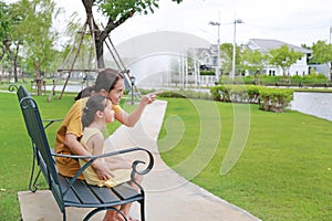 Portrait asian mother and daughter relaxing sitting on bench in garden outdoors. Mum pointing something with child girl looking in